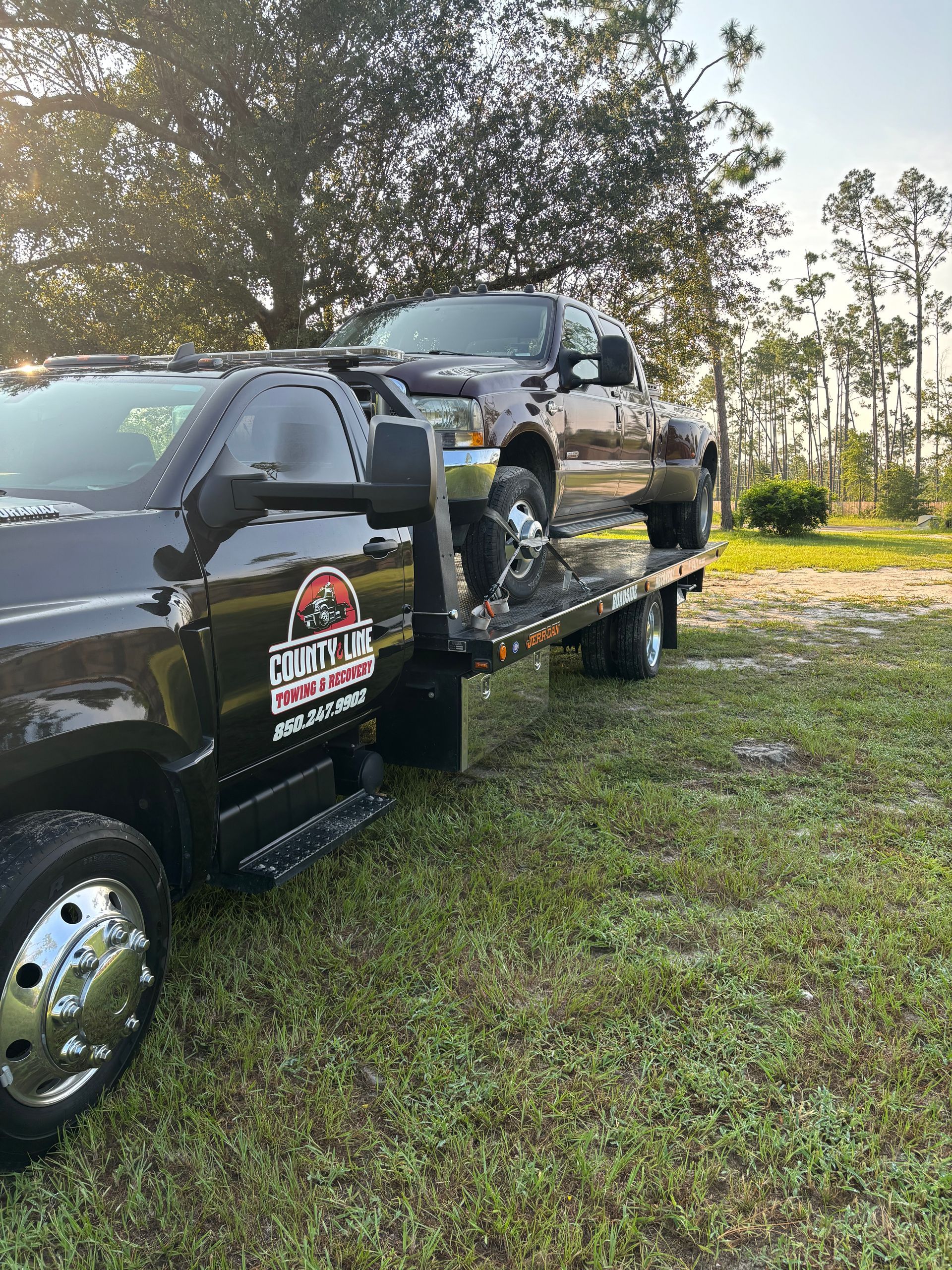 Tow truck hauling a dark-colored pickup truck in a grassy field with trees.