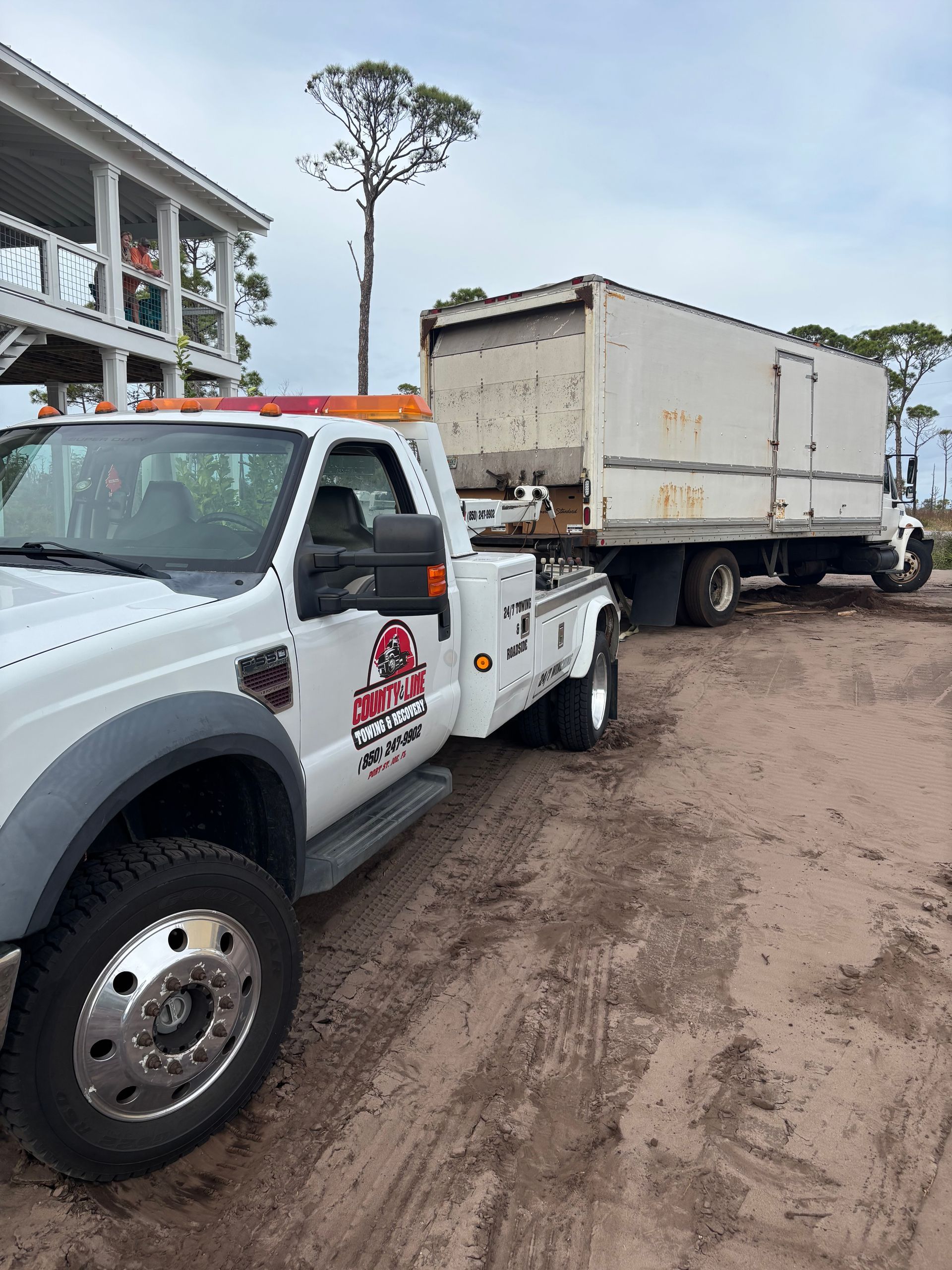 Tow truck towing a large white box truck on a sandy, dirt area near a house under a cloudy sky.