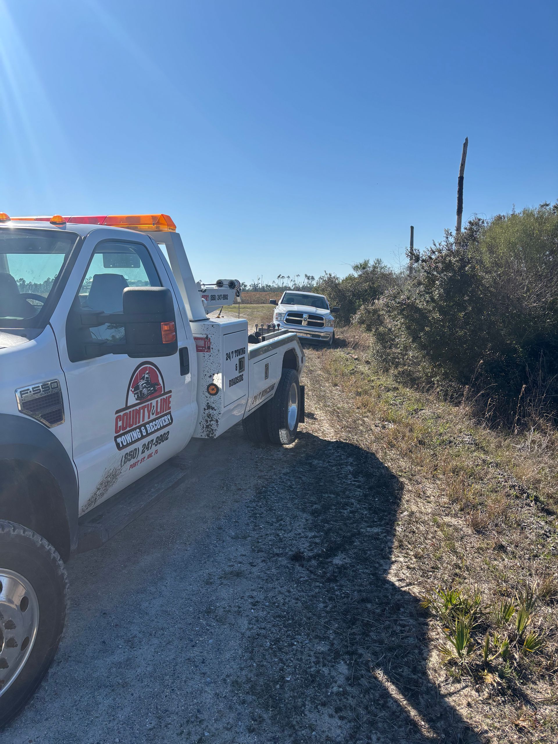Tow truck on a dirt road; white truck being towed. Bright blue sky and sunny outdoors.