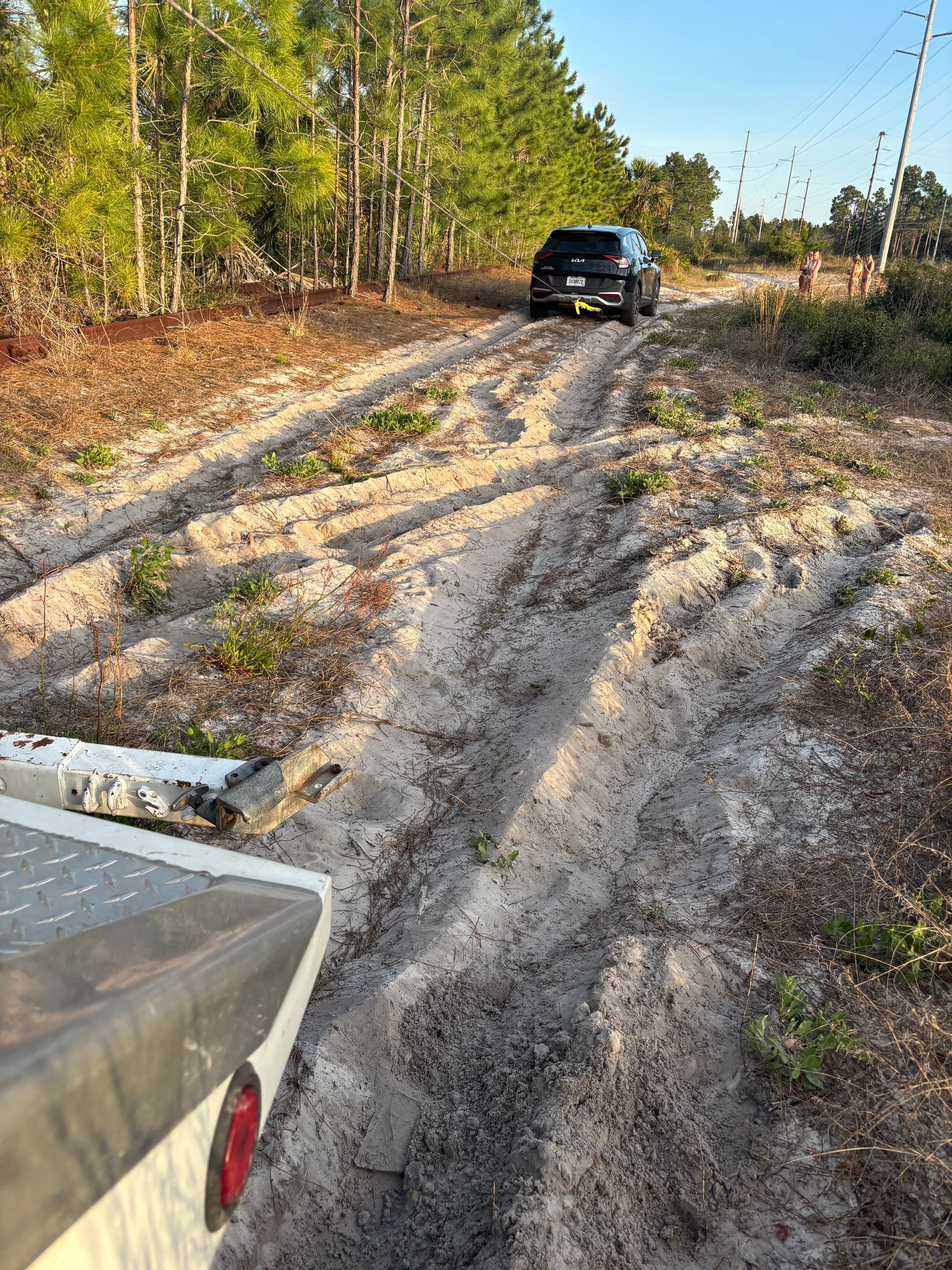 Truck towing a dark SUV on a sandy, dirt road through a wooded area.