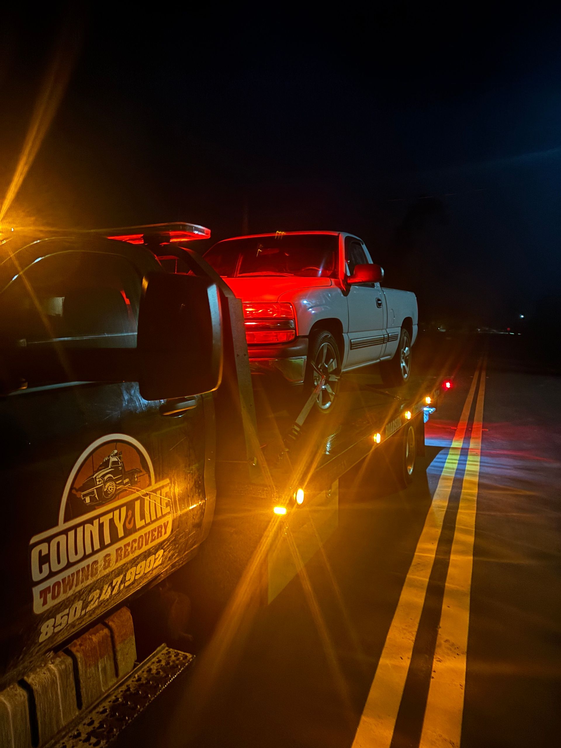 Tow truck hauling a white pickup truck at night on a highway. Orange and red lights are flashing.