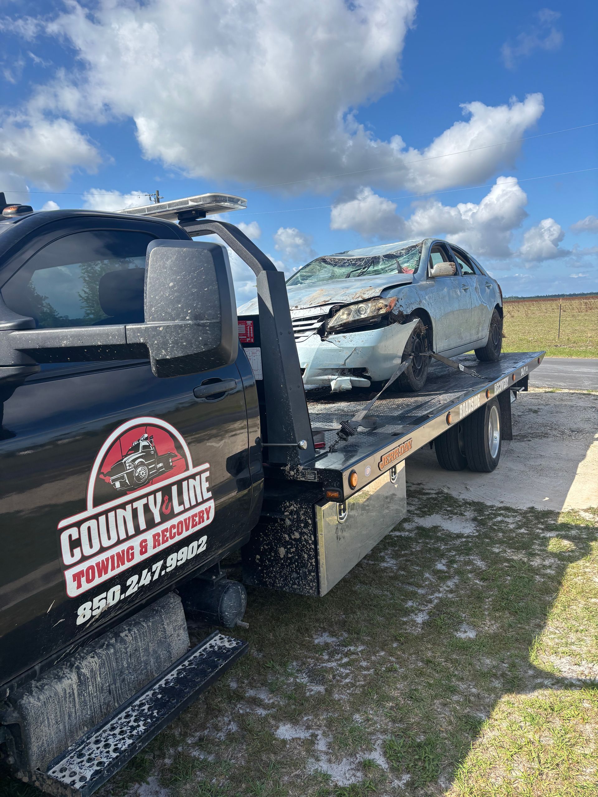 Tow truck hauling a damaged blue car. Sky and field in background. 