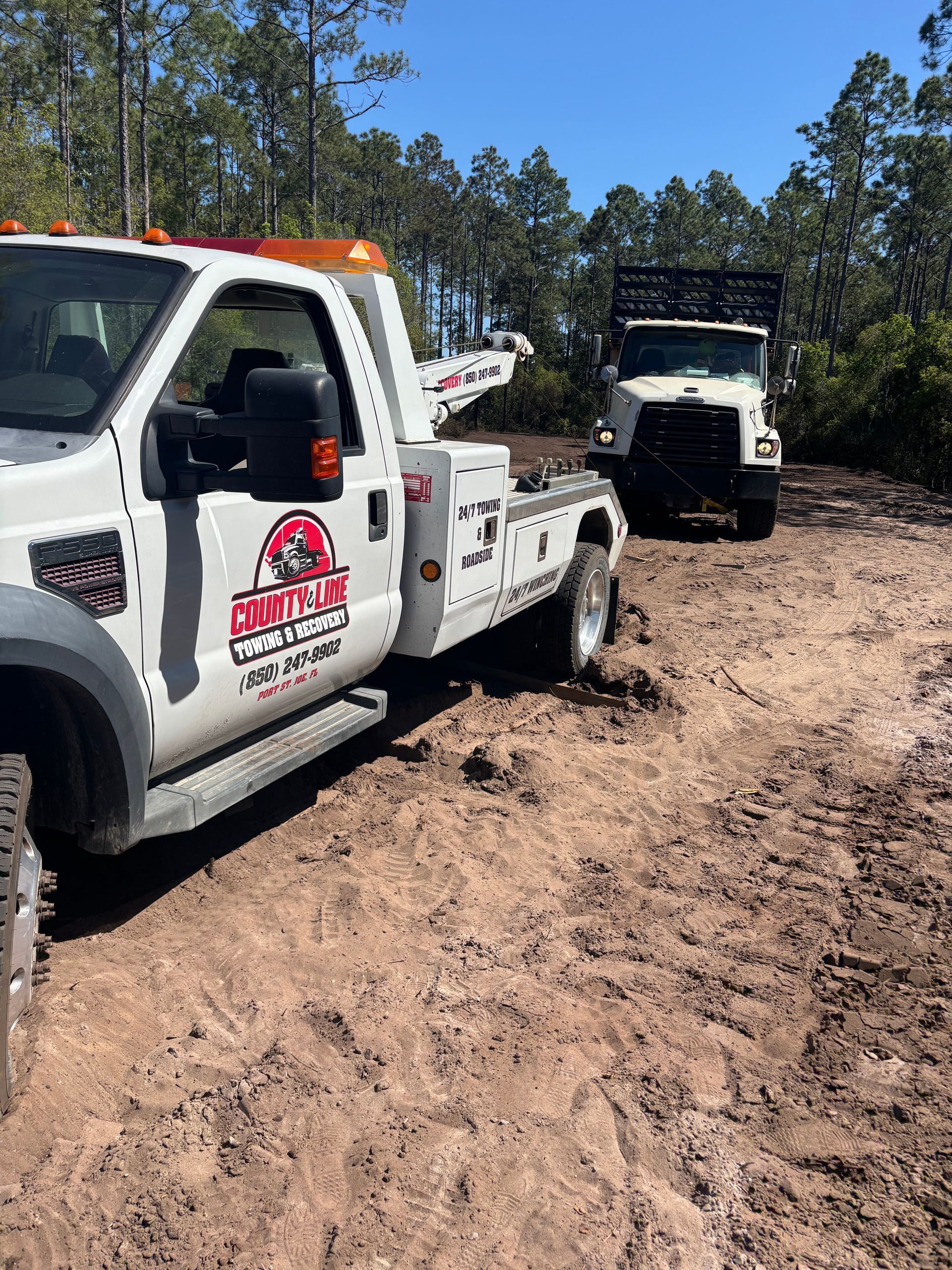 Tow truck towing a work truck on a muddy road. White trucks, red logo. Sunny day in a wooded area.