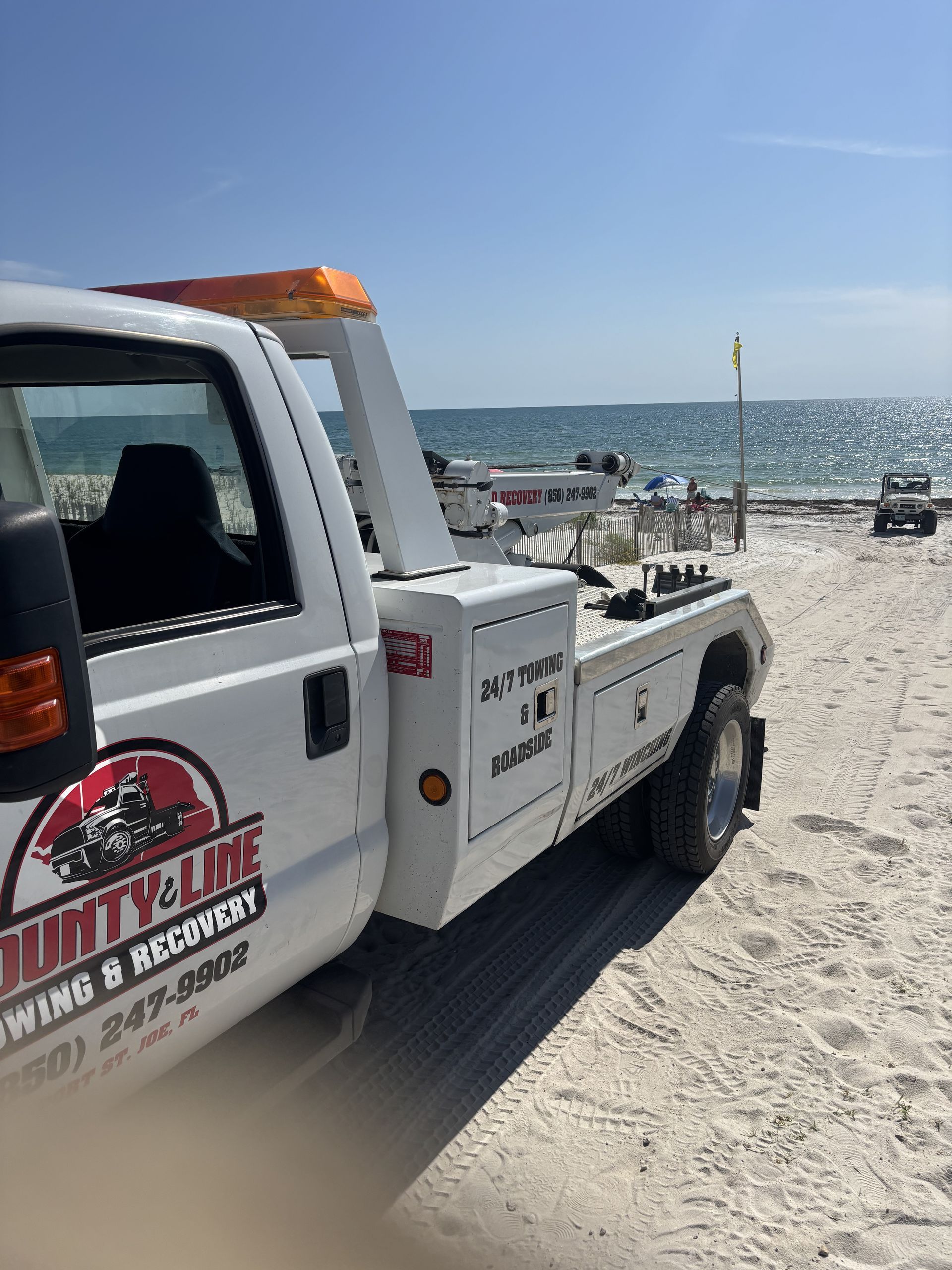White tow truck on a sandy beach with the ocean in the background.