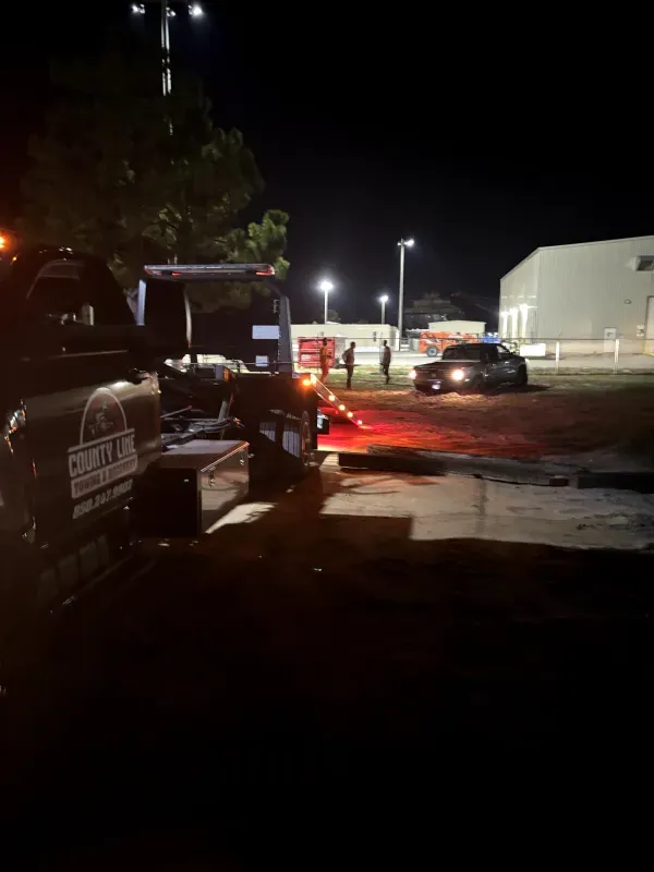 A tow truck is towing a car in a parking lot at night.