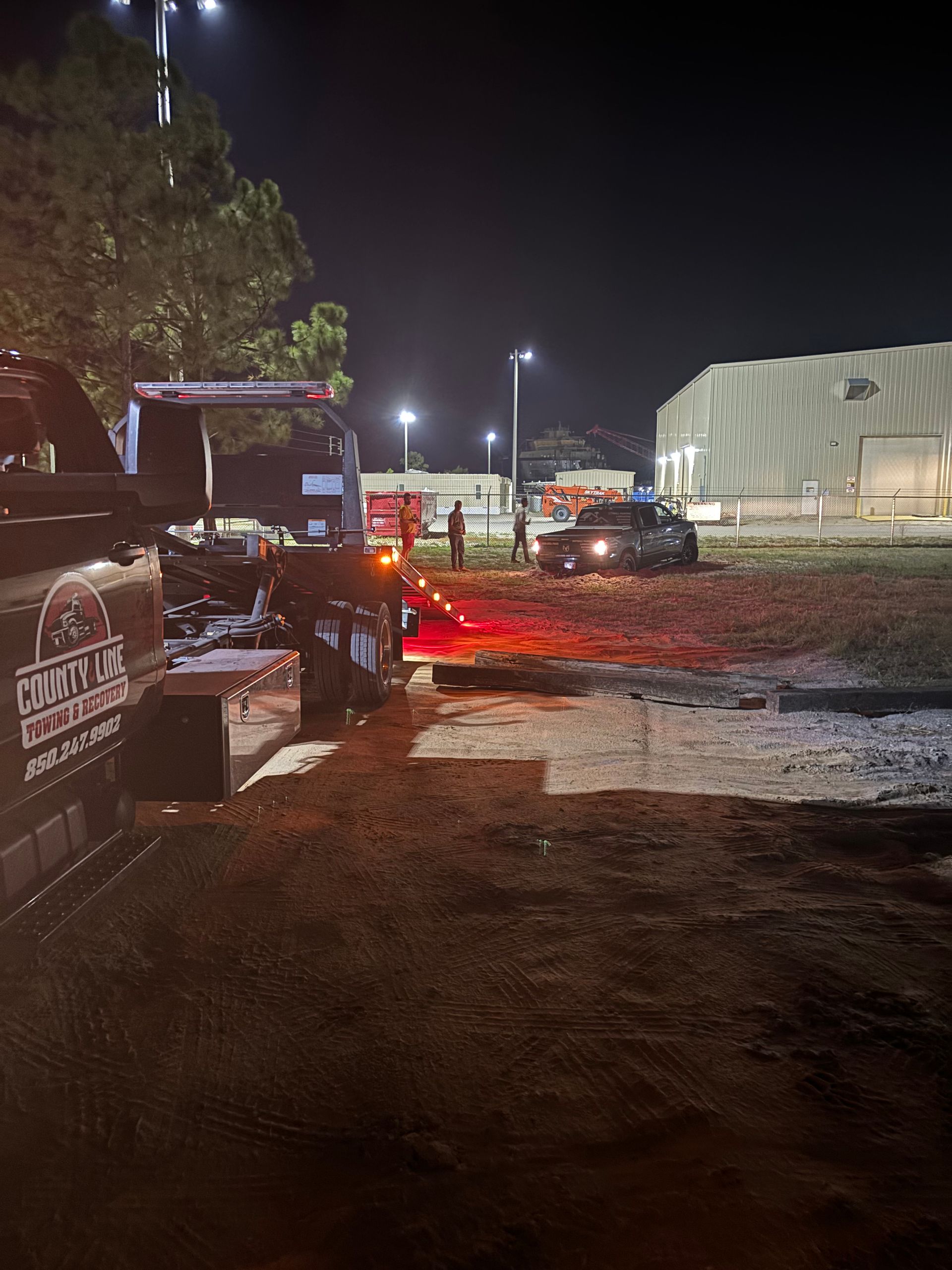 Tow truck pulling a vehicle at night. Red lights illuminate a dark field near buildings and a stadium.