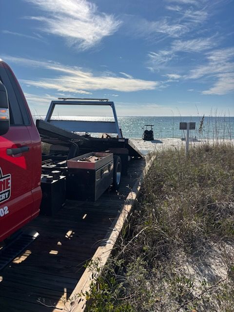 A red tow truck parked on a wooden boardwalk leading to a sunny beach with the ocean in the distance.