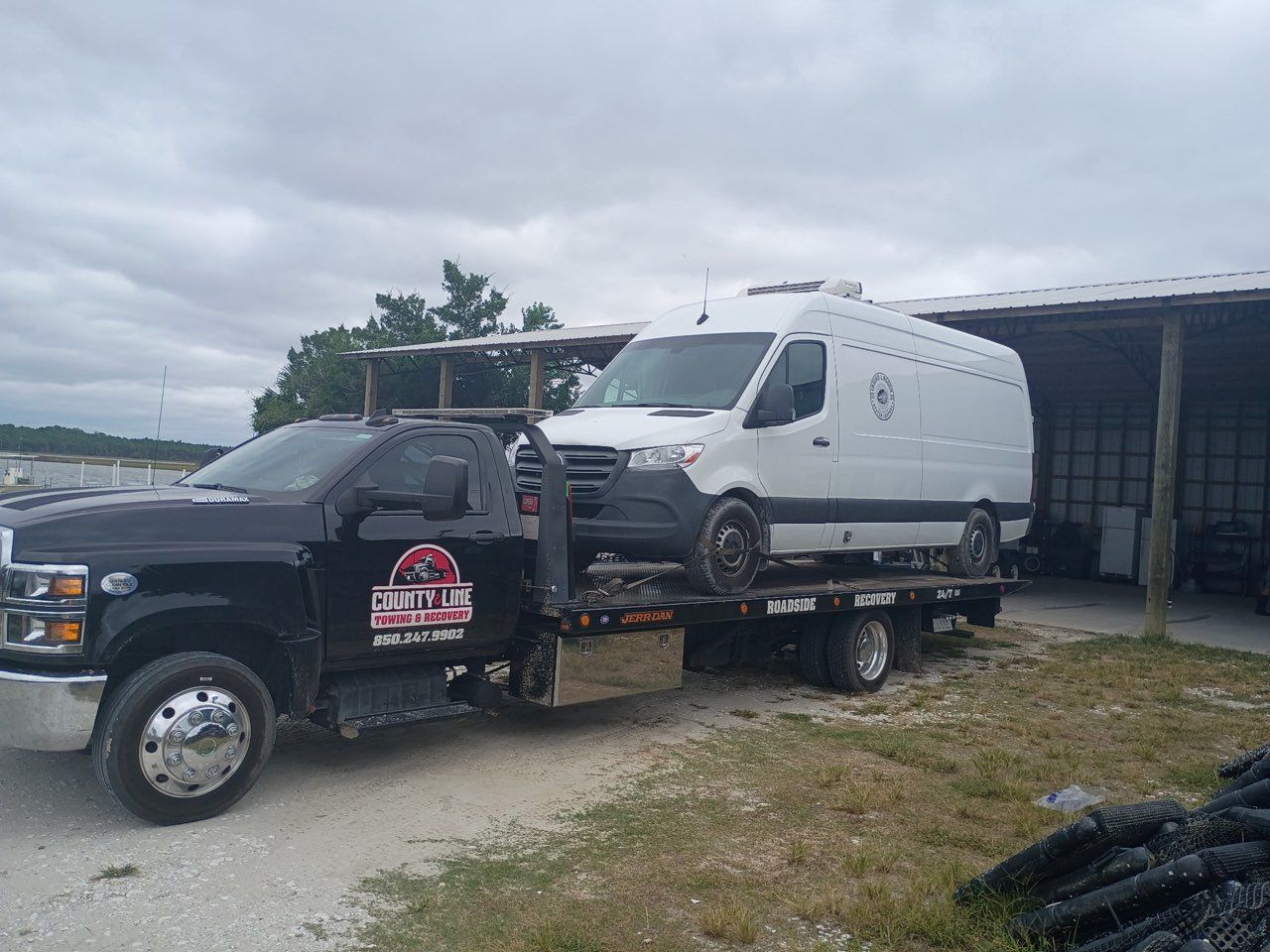 A black flatbed tow truck carrying a white cargo van parked near a shed on a gravel lot under a cloudy sky.