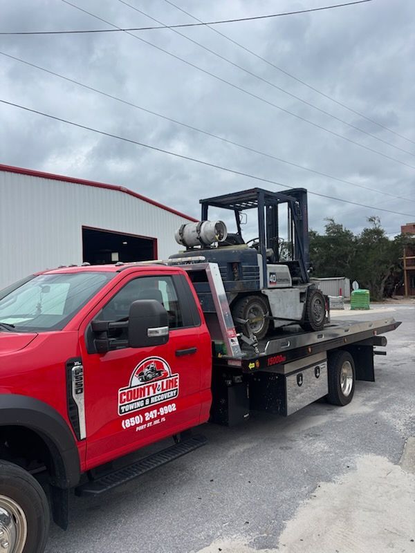 A red County Line Towing truck carrying a forklift on its flatbed in front of a white building under a cloudy sky.