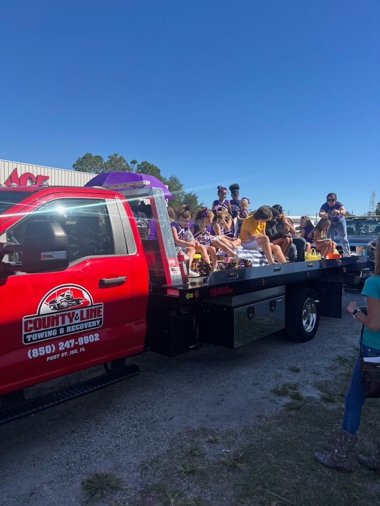 A red flatbed truck decorated as a parade float carries several people sitting on the back against a clear blue sky.