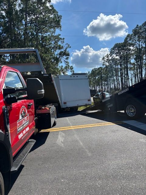 A red tow truck is parked on an asphalt road next to a white trailer being loaded onto a black flatbed trailer.