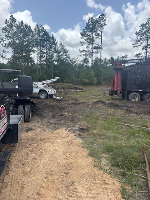 A tow truck pulls a white vehicle stuck in a muddy, rural clearing near a large industrial wood chipper.