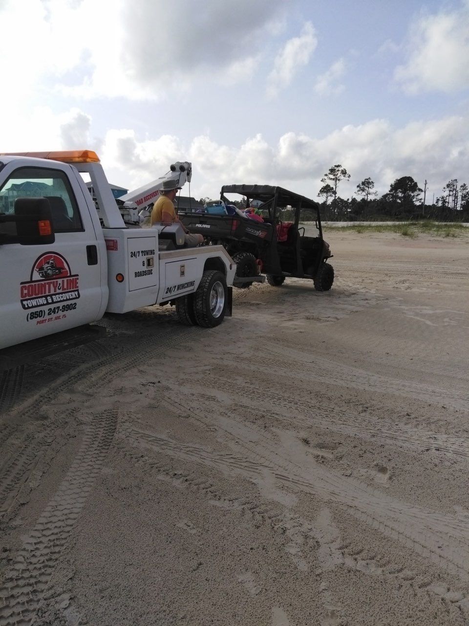 A white tow truck is parked on a sandy beach, preparing to tow a dark-colored utility vehicle.