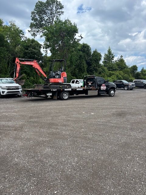 An orange mini-excavator loaded on a black flatbed tow truck parked in a gravel lot with other vehicles and trees nearby.
