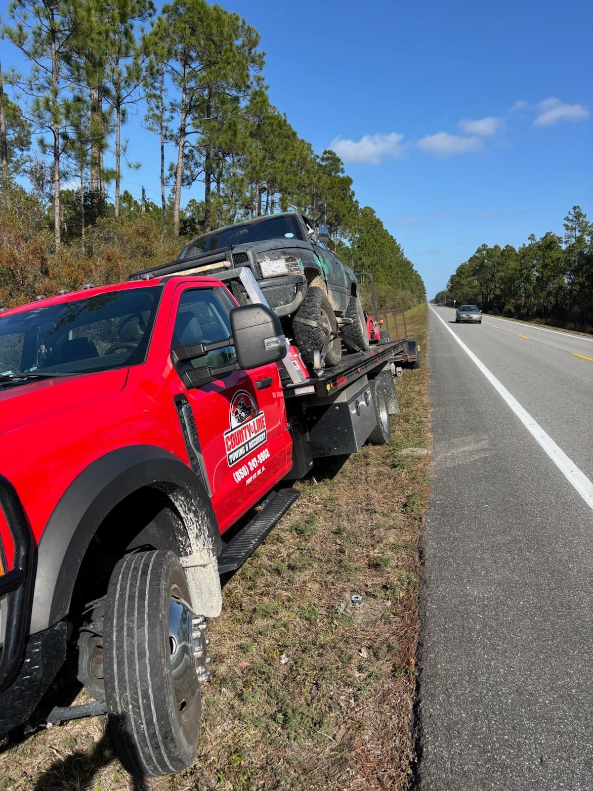 A red flatbed tow truck parked on the grassy shoulder of a sunny highway, loaded with a dark-colored vehicle.