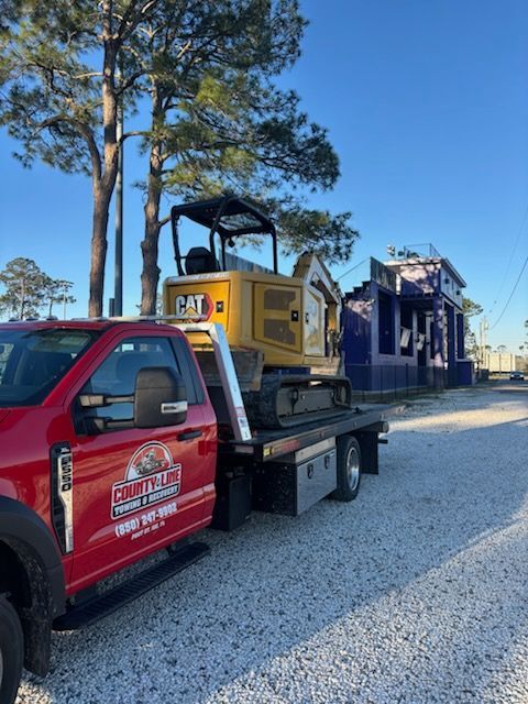 A yellow Caterpillar mini-excavator loaded on a red County Line flatbed truck parked on a gravel lot near a building.