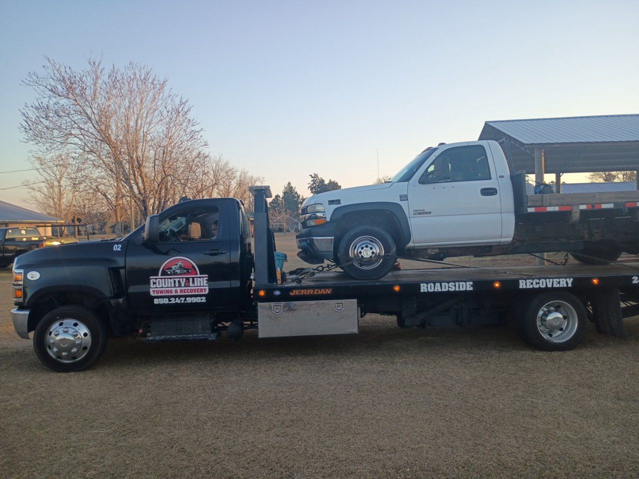 A black tow truck transporting a white pickup truck on a gravel lot during a sunny day.