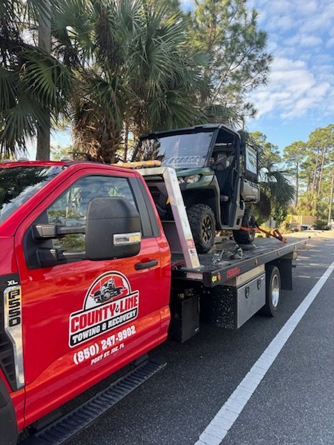A red County Line Towing truck carrying a green side-by-side utility vehicle on its flatbed on a sunny day.