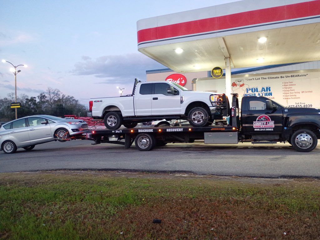 A white pickup truck sits on a flatbed tow truck, which is towing a silver sedan at a gas station.
