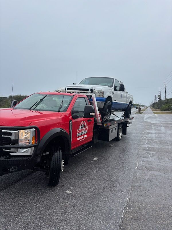 A bright red tow truck carrying a white pickup truck on its flatbed on a paved road under an overcast sky.