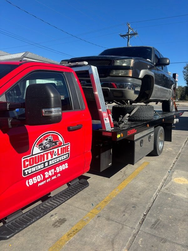 A red County Line Towing truck carrying a black pickup truck on a flatbed trailer parked on a concrete lot.