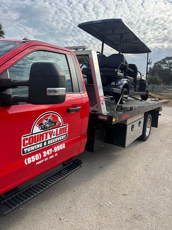 A red County Line Towing and Recovery flatbed truck carrying a black golf cart under a cloudy sky.