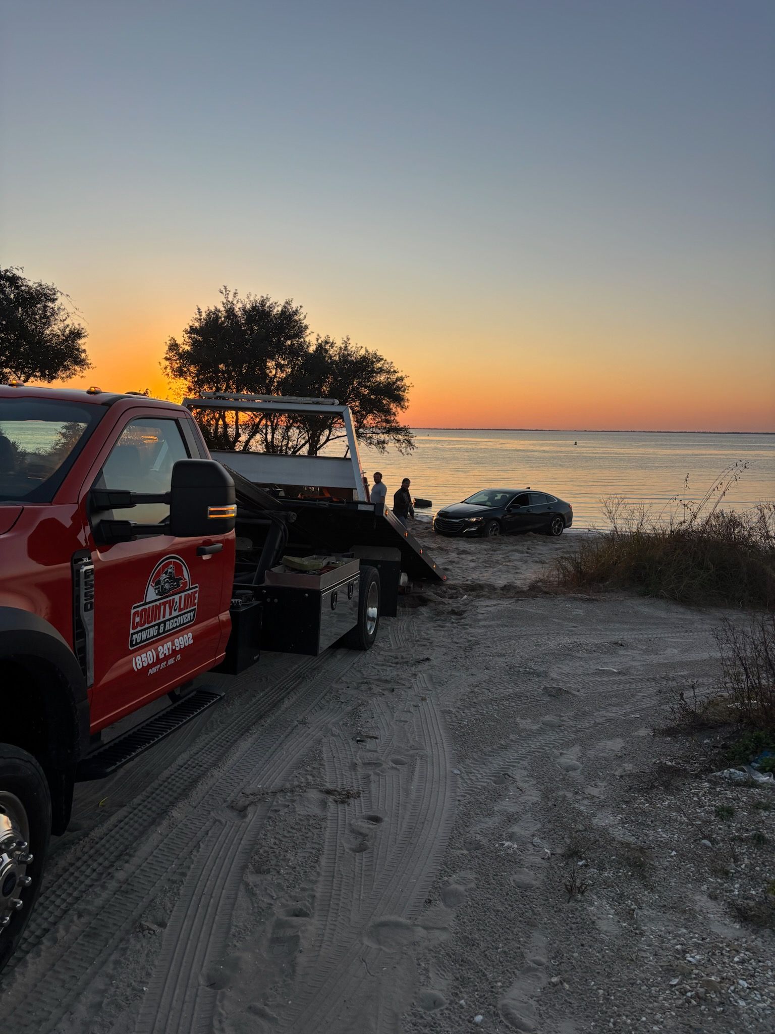 A red tow truck parked on a sandy beach at sunset, preparing to recover a dark sedan stuck near the water’s edge.
