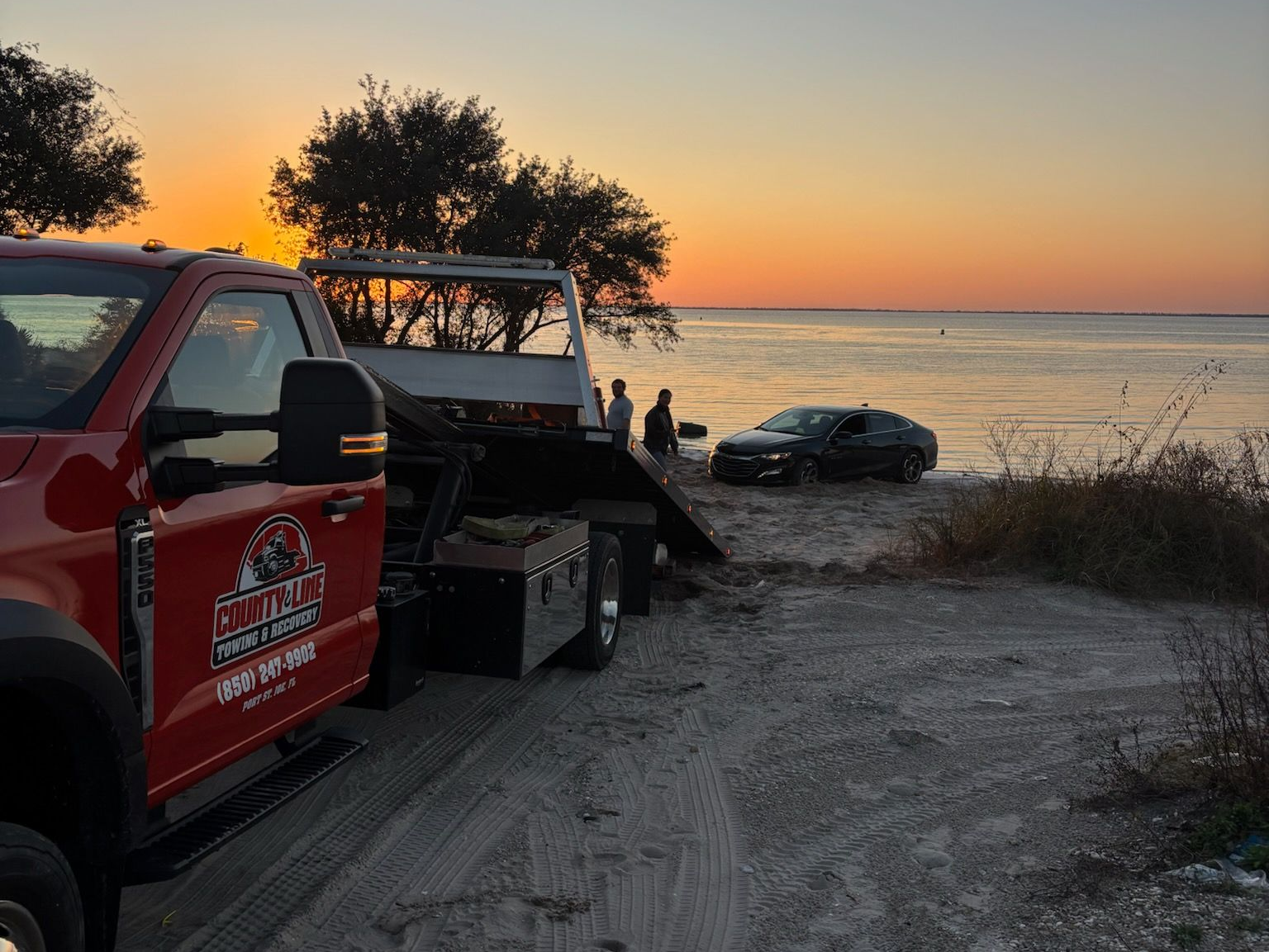 Tow truck and red pickup by a lakeshore at sunset, with a car pulled onto the roadside nearby