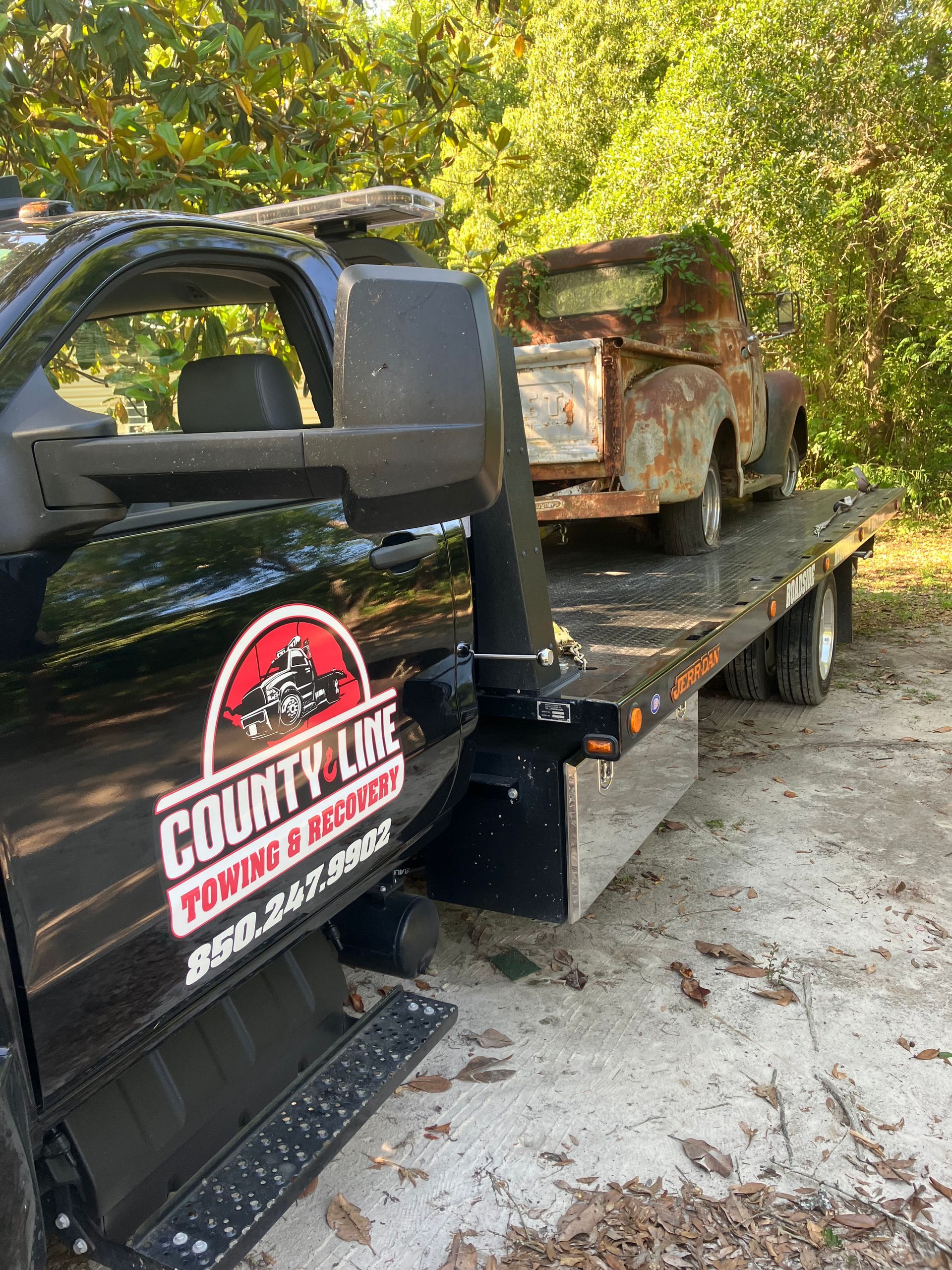 Tow truck hauling a rusty vintage truck. County Line Towing logo on the truck's door. Outdoors, trees in the background.