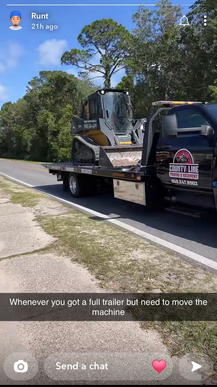 Tow truck carrying a skid steer loader on a flatbed trailer, on a road, trees in background.