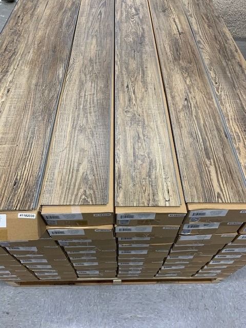 Stacked boxes of wood-look flooring planks with gray-brown grain patterns in a store aisle.