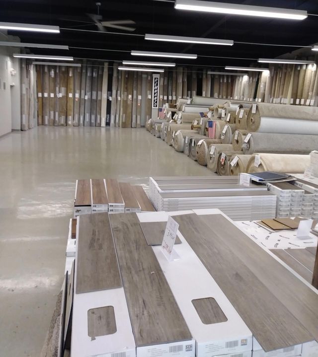 Furniture showroom with stacked wood-look panels and rolls of flooring in a large empty warehouse-like space