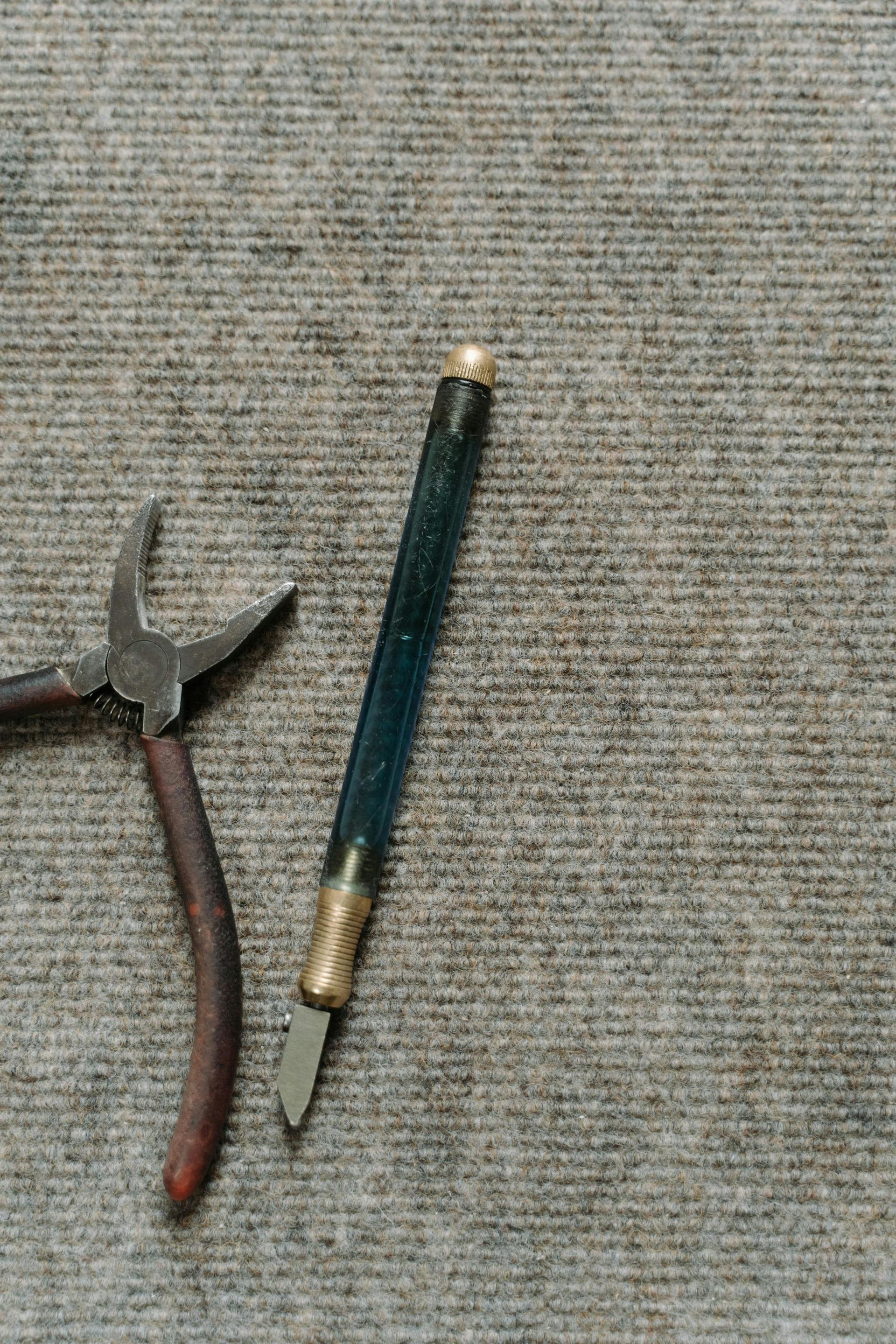 Pliers and a dark blue pen-like tool on a gray fabric surface