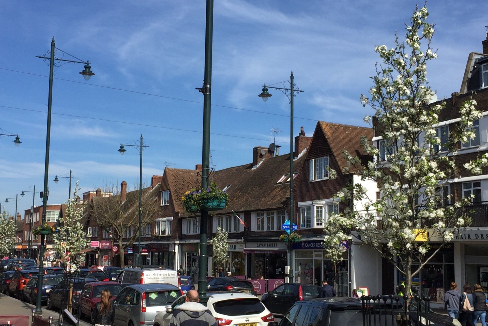 Busy street with parked cars, pedestrians, and brick shops under a blue sky with blooming trees