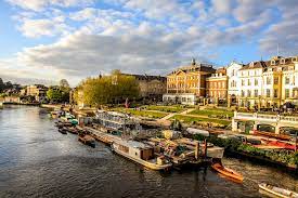 Riverside cityscape with moored boats, a tree-lined promenade, and colorful historic buildings in warm sunlight