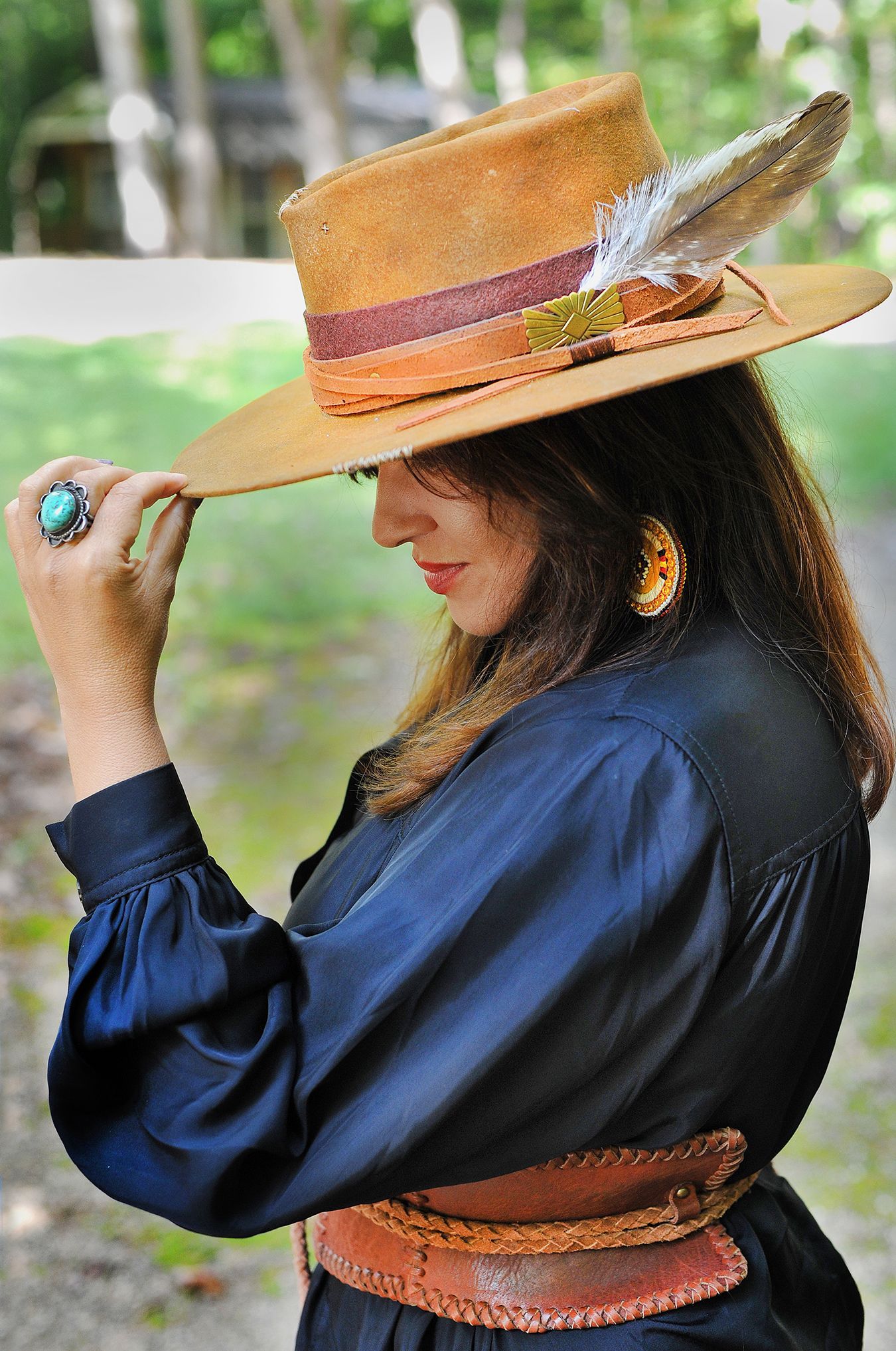 A person wears a brown leather jacket, green shirt, and patterned poncho with a fedora, posed against a green background.