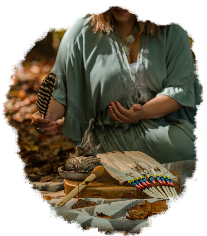 Woman holding a feather, surrounded by ritual items: bowl, drum, fan. Soft light, outdoor setting.