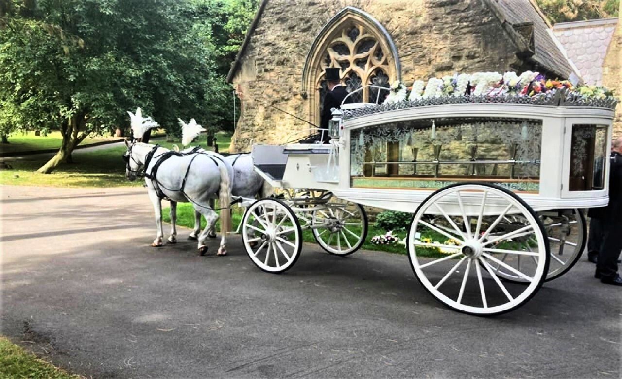 White horse-drawn hearse with a floral arrangement, in front of a stone funeral home.