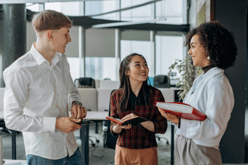 Drie collega's voeren een gesprek in een kantooromgeving, met notitieboekjes in de hand en een neutrale gezichtsuitdrukking.