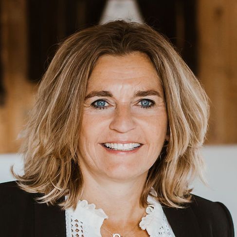 Vrouw met blond haar lacht, draagt een zwarte blazer en een witte blouse, binnen.