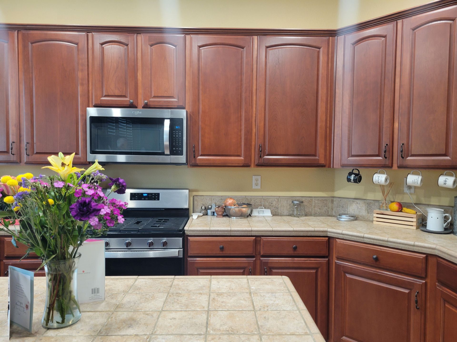 Kitchen with wooden cabinets, stainless steel appliances, beige tile counters, and a vase of purple flowers.
