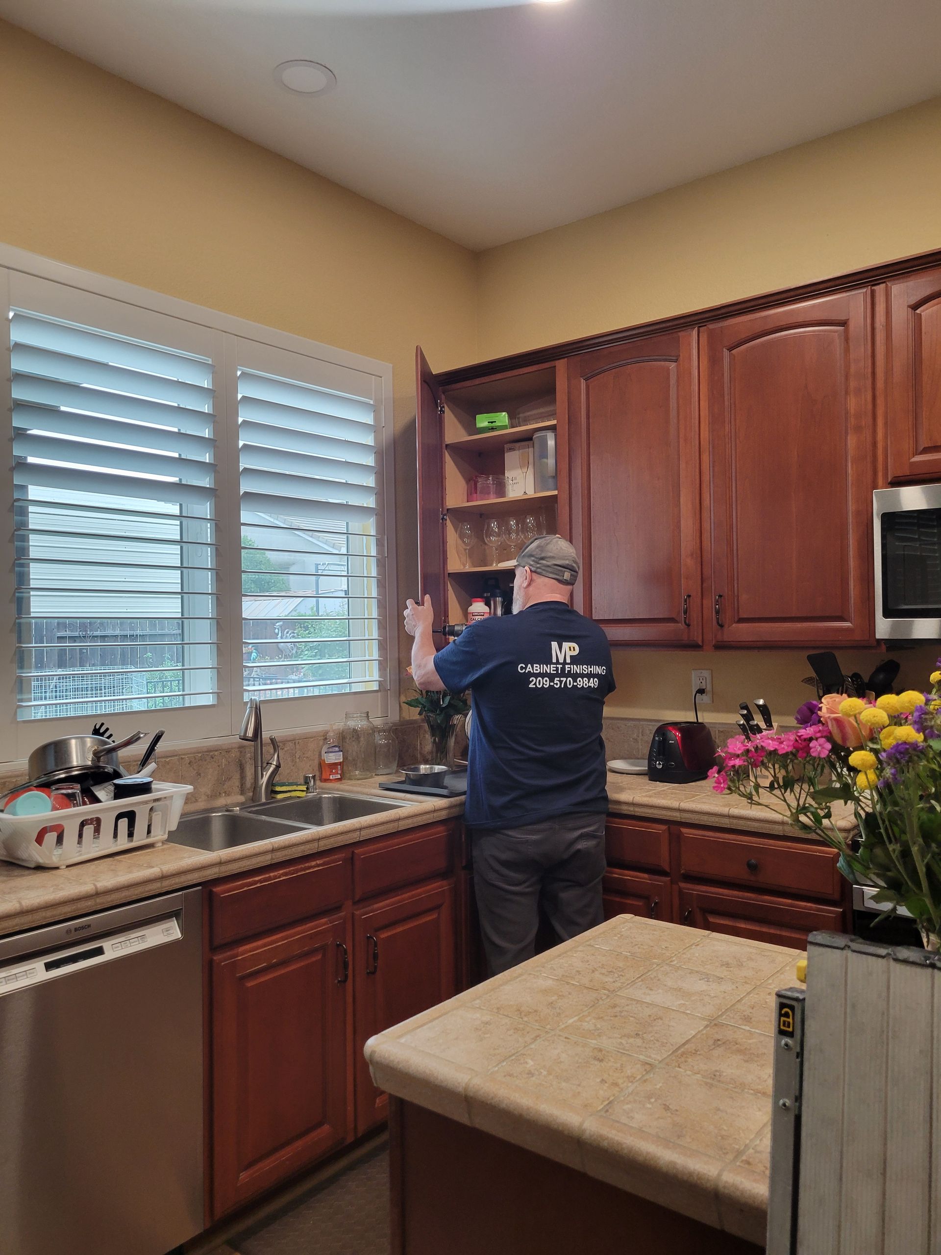 Person washing dishes at a kitchen sink beside a window, with wood cabinets and a flower arrangement.