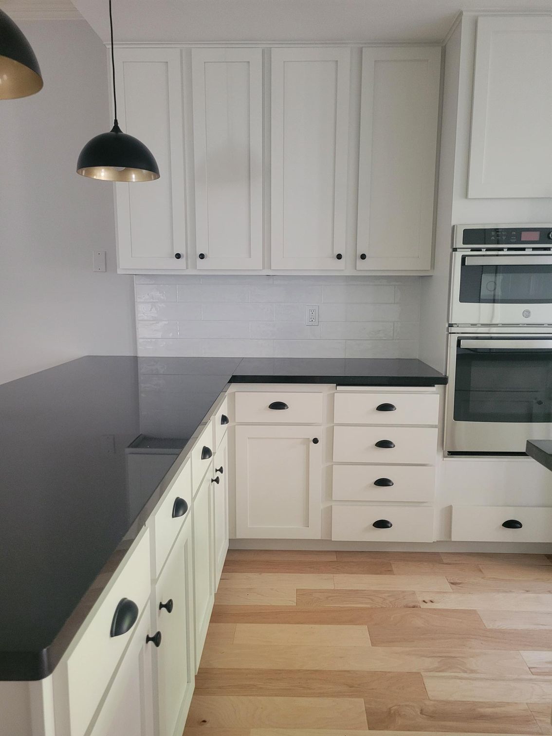 Modern kitchen with white cabinets, black countertop, pendant light, built-in oven, and light wood floor
