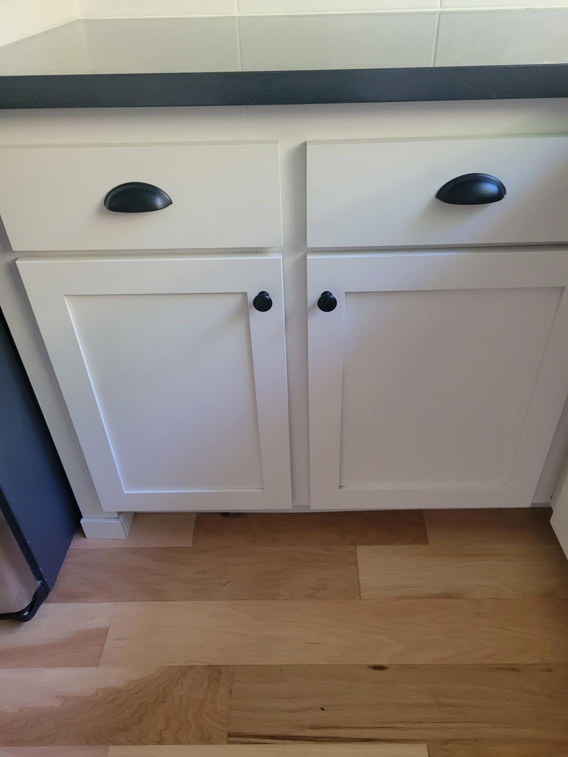 White kitchen cabinet with black knobs under a countertop on a wood floor.