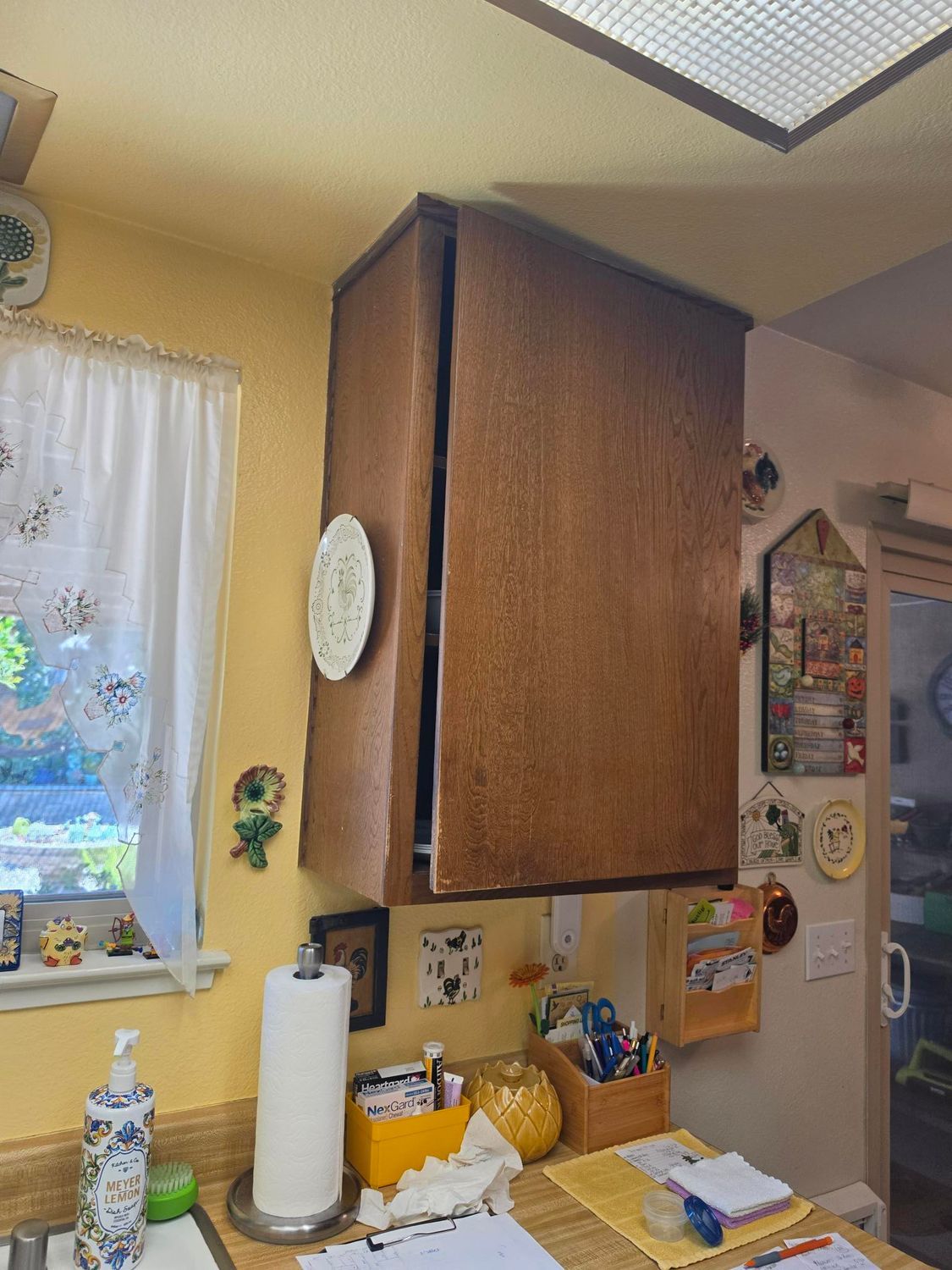 Kitchen corner with wooden wall cabinet, sink, paper towel roll, and window curtain.