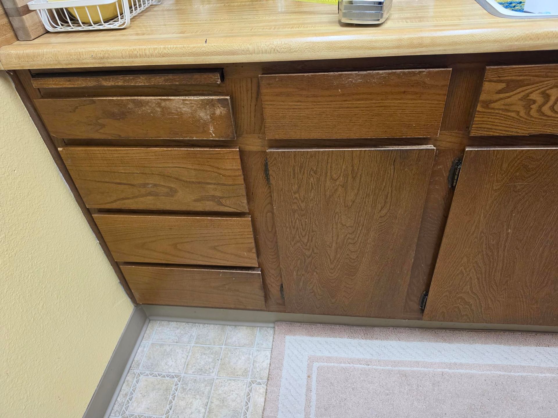 Wooden kitchen cabinet with drawers and doors beneath a countertop, next to a tiled floor and wall.