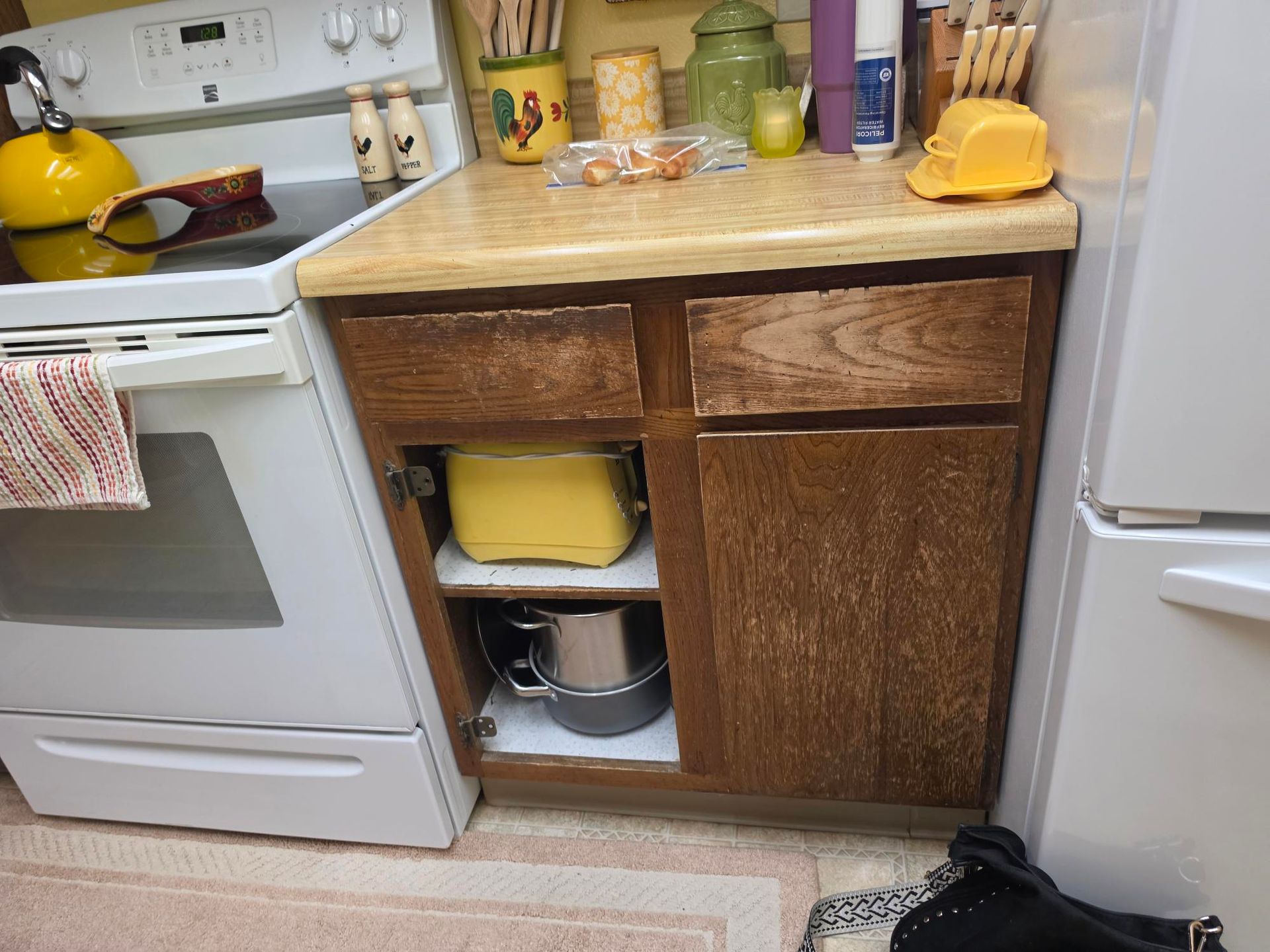 Kitchen corner with stove, worn wooden cabinet, countertop items, and refrigerator.