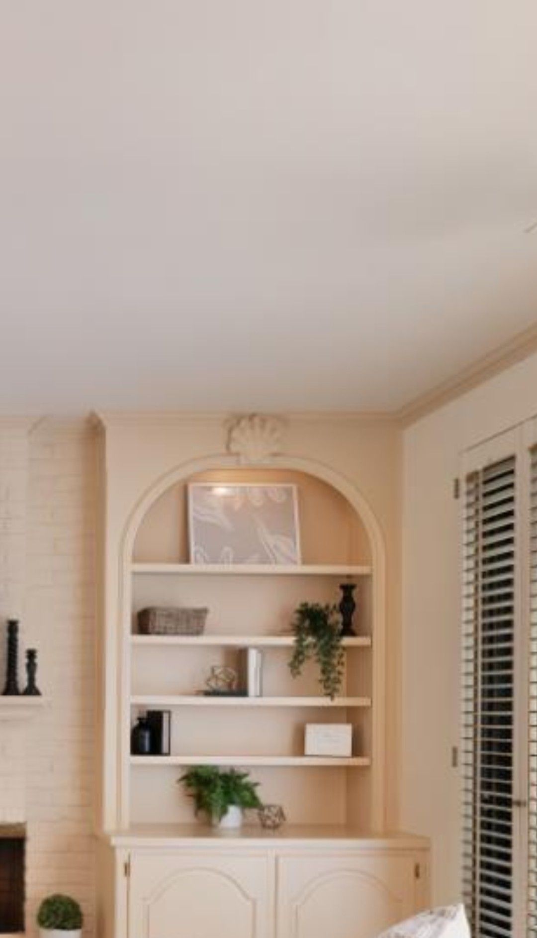 Beige arched built-in shelving with decor and plants in a bright room, beside a window with shutters