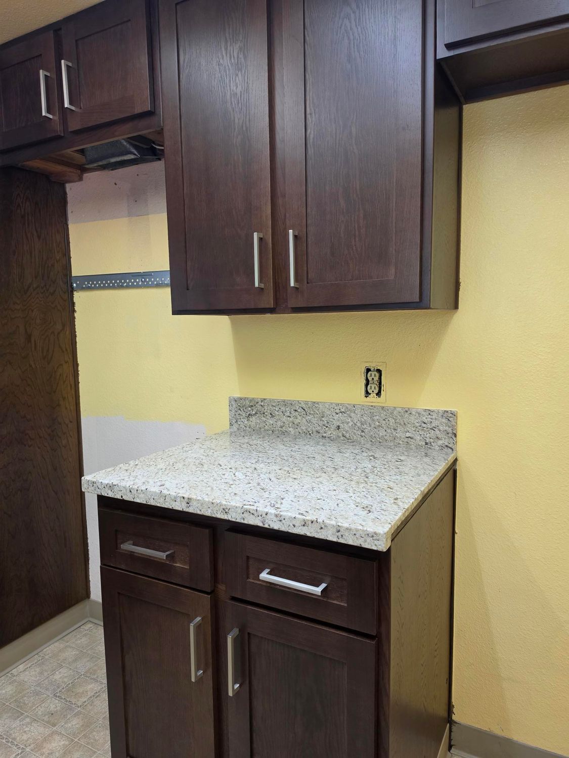 Small kitchen cabinet and granite countertop between dark wood cupboards, with a yellow wall behind it.