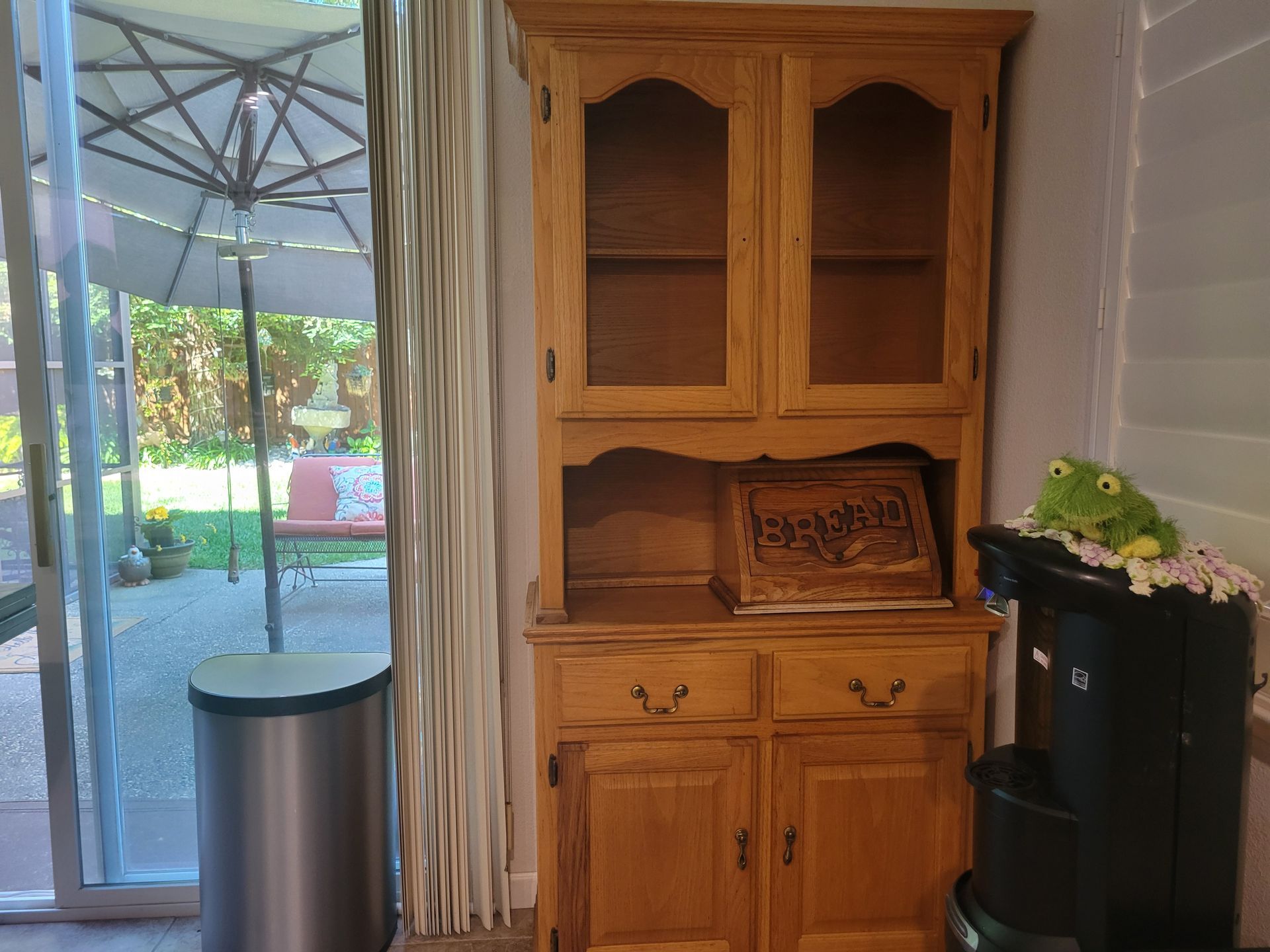 Wooden hutch cabinet beside sliding glass door, with a trash can and curtain in a bright room.